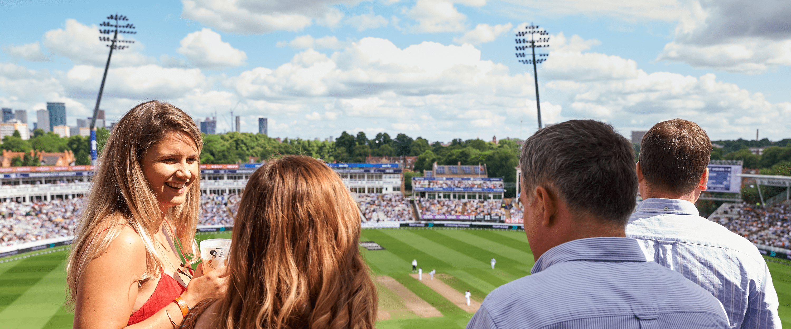 Guests stood chatting in the sunshine at Edgbaston Stadium. with a view of the cricket pitch and match in the background