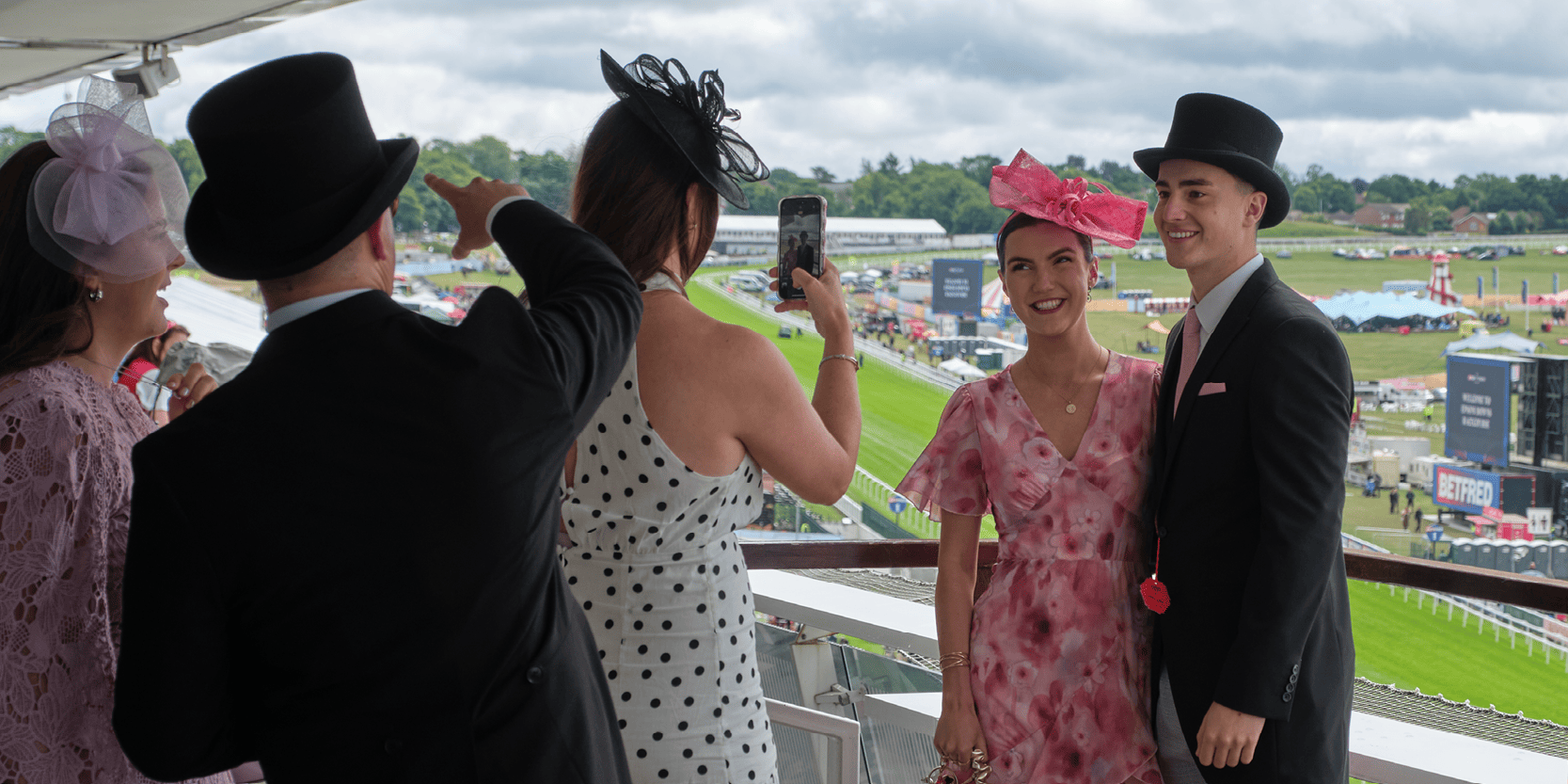 Guests smiling for a photo on the balcony of the hospitality suite at Epsom Downs Racecourse for the Epsom Derby Festival