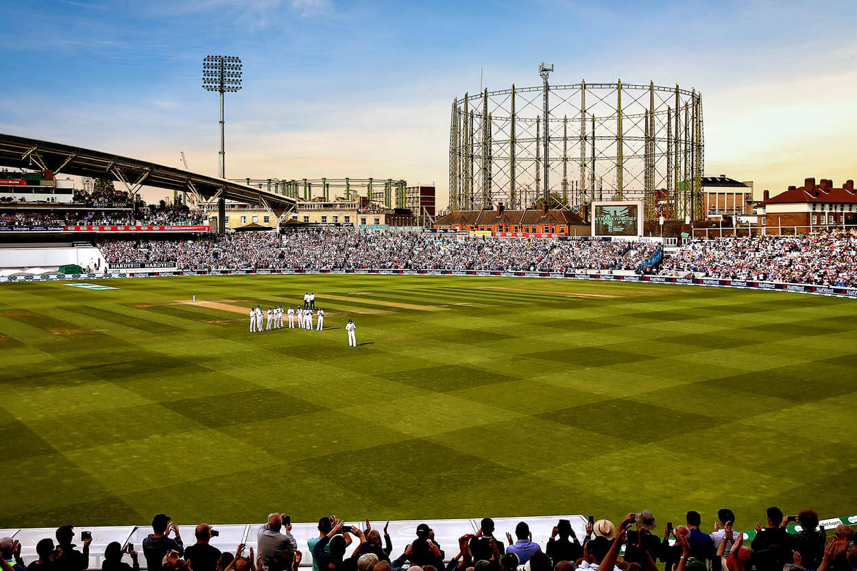 View of the cricket pitch at the Kia Oval cricket ground with England players gathered in a group for a team talk with views of the London skyline in the background