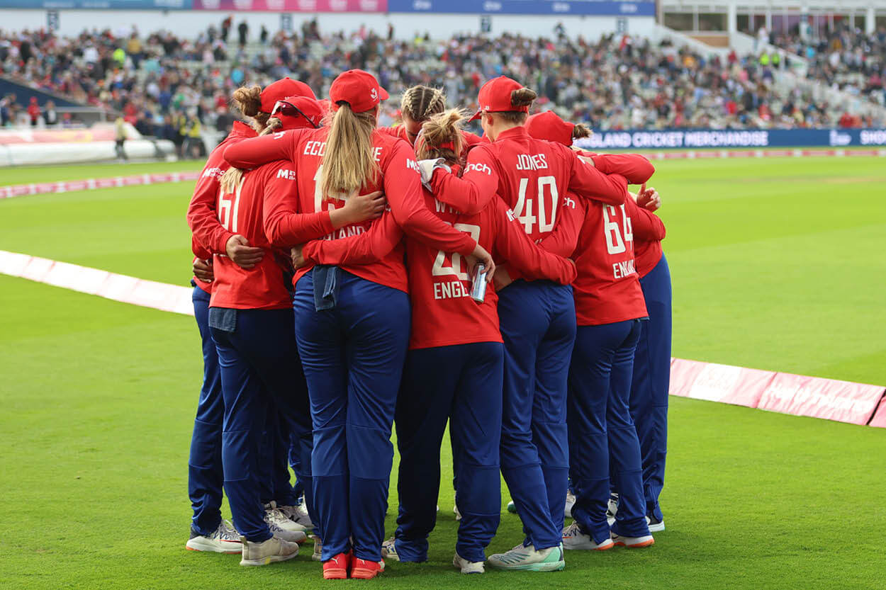 Women's England IT20 cricket team hugging in celebration on the pitch