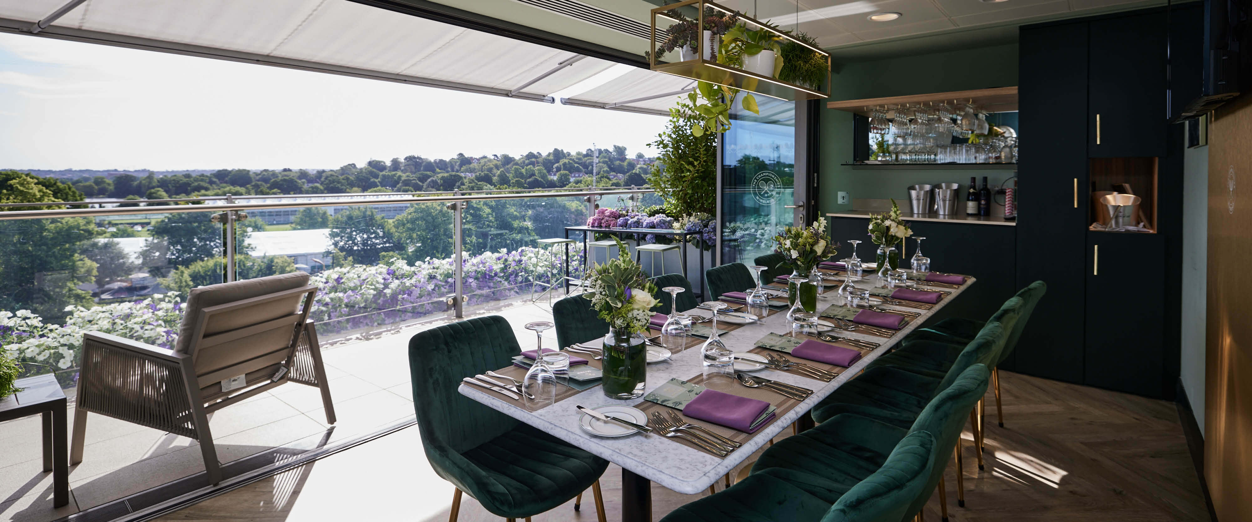 Skyview Suite dining area with opened terrace doors to private balcony with seating overlooking the courts and grounds at The Championships Wimbledon