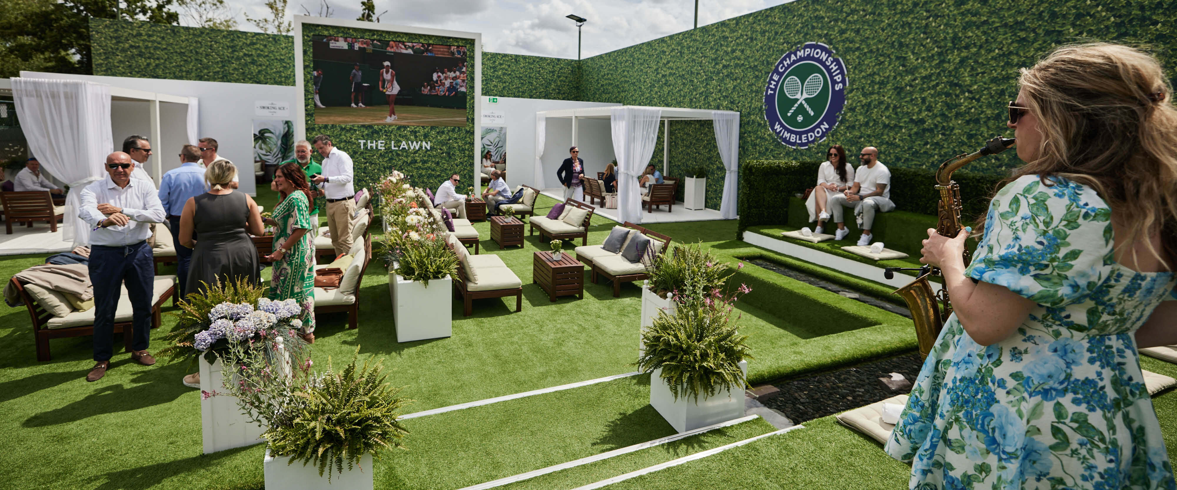 The Lawn English garden area with saxophonist playing and guests sitting on sofas enjoying drinks at The Championships Wimbledon